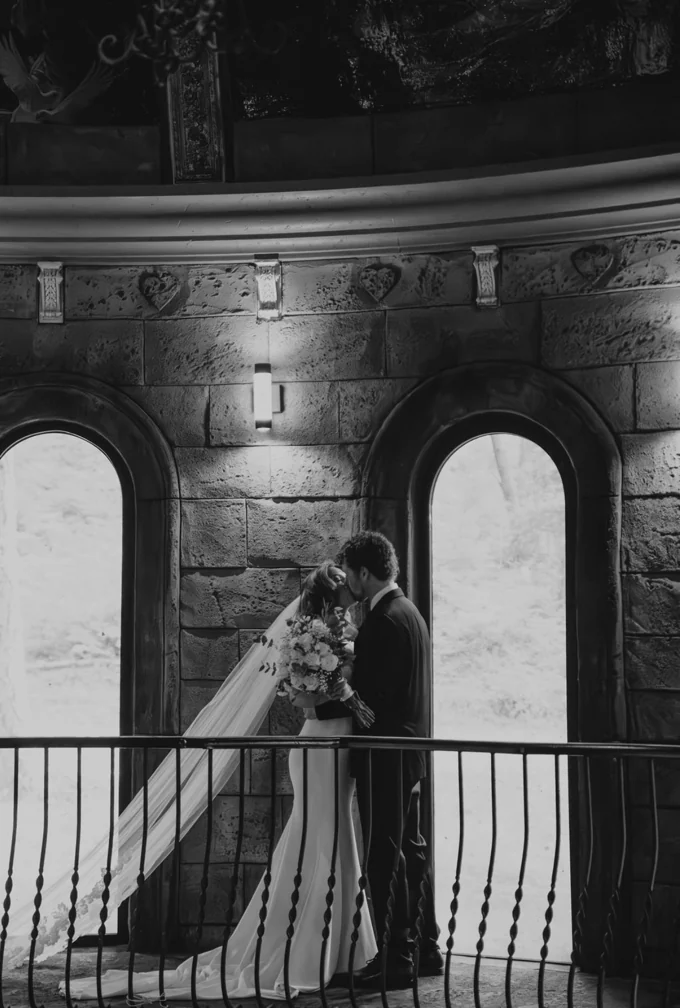 Black and white portrait of newlyweds kissing beside the arched chapel windows