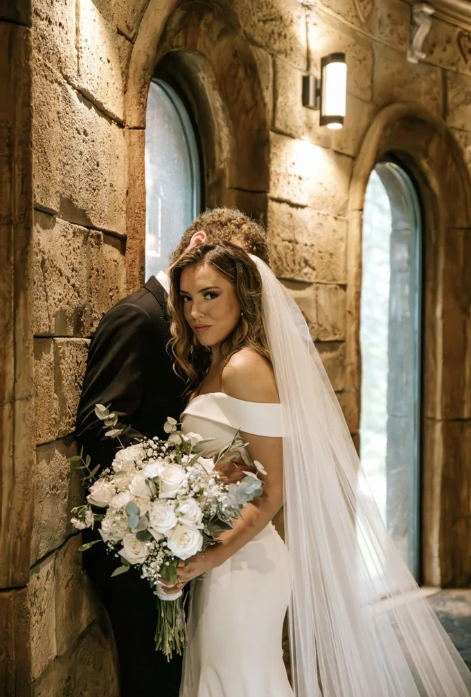 Bride holding a white bouquet beside the chapel windows with the groom behind her