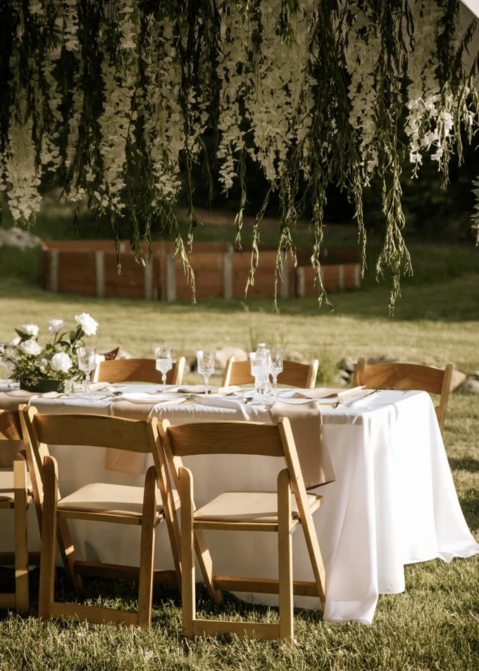 White linen table setting with wooden chairs beneath hanging white flowers