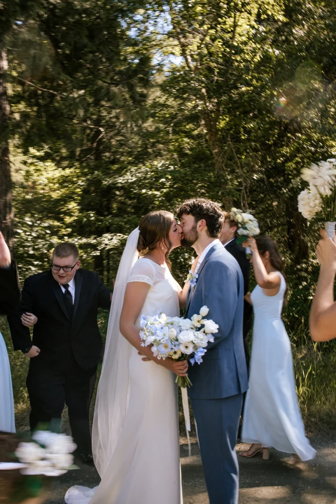 Newlyweds kissing outdoors with their wedding party nearby