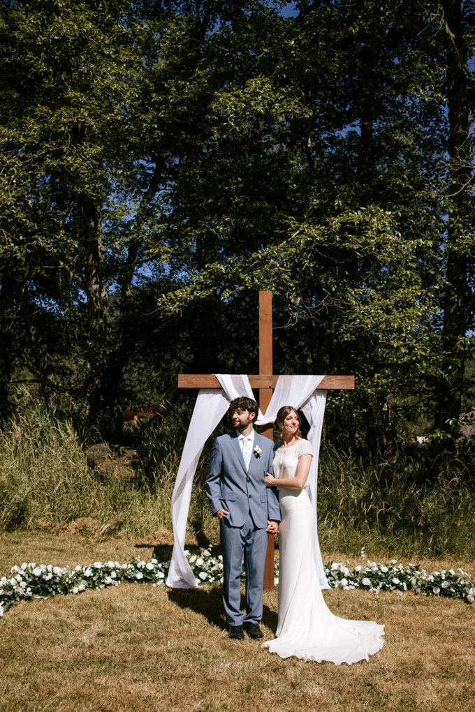 Newlyweds standing before a wooden cross at an outdoor ceremony